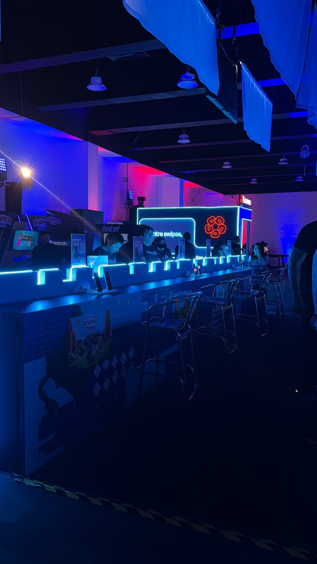 Pasillo de estaciones de juego con luces de neón azules y rojas en el Banamex Super Indie Games 2026.